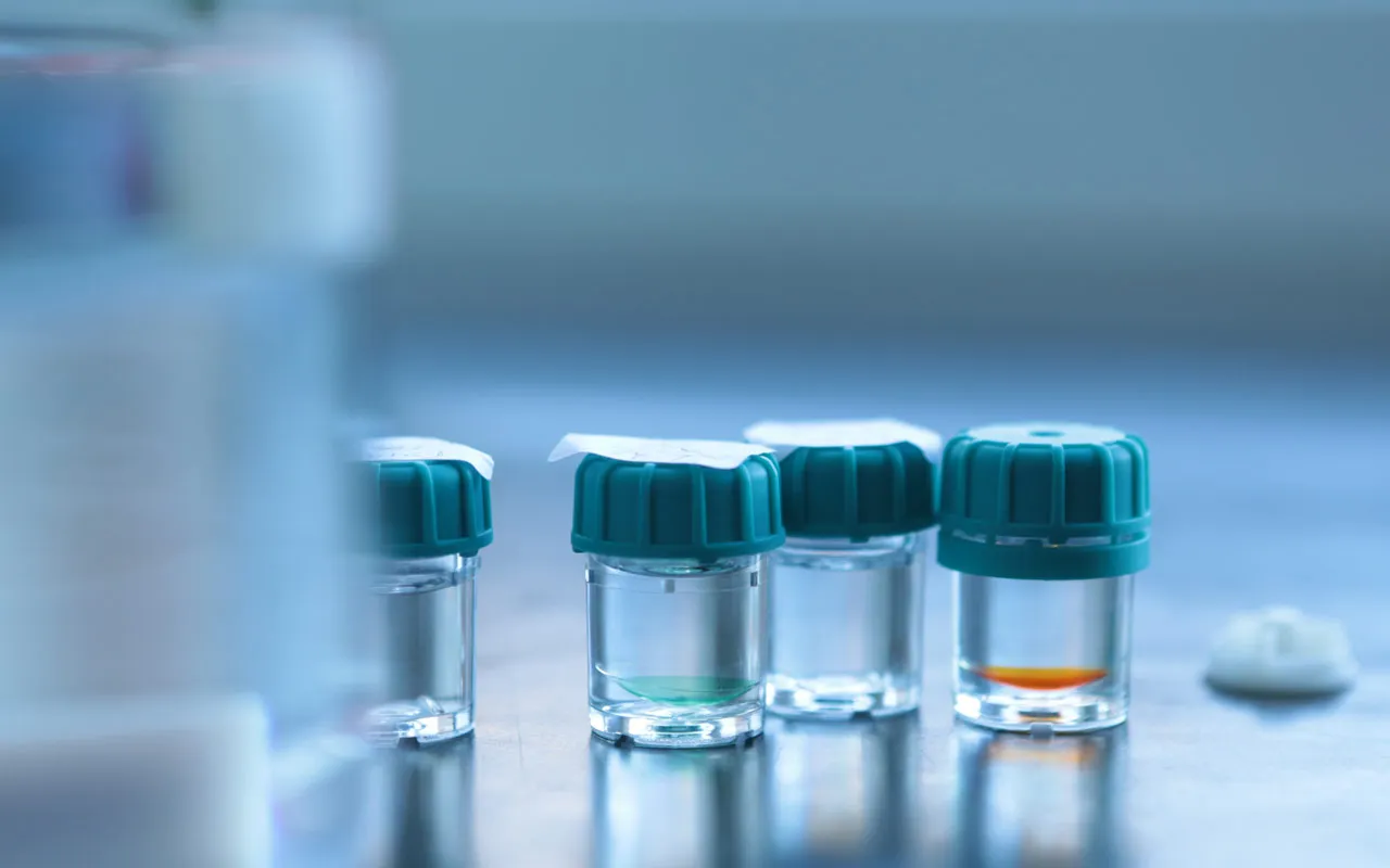 Štiri prozorne stekleničke s kontaktnimi lečami v raztopini, s turkiznimi pokrovčki, na modri laboratorijski površini